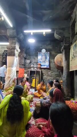 This video shows a group of women sitting in a circle on the floor of what appears to be a temple or shrine. They are all dressed in traditional Indian clothing. Some of the women are clapping their hands, while others appear to be singing or chanting. There is a framed picture of a deity on the wall behind them, and various religious artifacts are visible. The lighting is dim, with overhead fluorescent lights illuminating the scene. The architecture suggests an old stone building, with rough-hewn pillars and walls. One woman in the foreground is wearing a bright green outfit, and her back is to the camera. The overall atmosphere seems to be one of prayer or devotion.