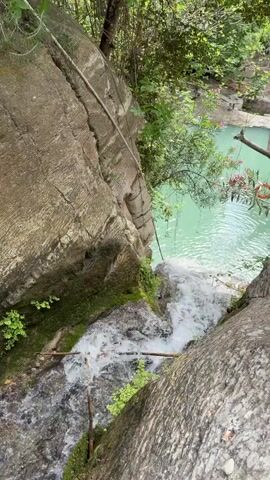 The camera is angled downward toward a small waterfall cascading from a higher pool of bright turquoise water. The immediate foreground consists of two large, striated rock formations, one to the left and one to the right of the frame. The waterfall flows between these rocks, surrounded by lush green moss and small plants. The higher pool is partially visible in the background, framed by verdant trees and foliage. A long, weathered rope hangs down from the top of the rock on the left, possibly for support. The overall setting is a natural, wooded area.