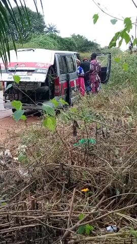 The video captures a white and red commercial passenger van or minibus, labeled "THE TRANSPORTERS" and "Flight," stationary on a dirt road in a rural area of Okele, Nigeria. The vehicle appears to have sustained damage to its rear, with the bumper either detached or heavily damaged, revealing internal components. Both the rear door and a side door are open.

Several individuals are gathered around the van. A man wearing a grey hoodie and dark trousers stands at the rear of the vehicle, seemingly inspecting or attending to something. Three other people, including two women dressed in colorful patterned traditional attire, are congregated near the open side door, possibly entering, exiting, or conversing.

The setting is characterized by an unpaved, possibly muddy, road flanked by dense, overgrown vegetation, including tall grasses, bushes, and trees. Palm trees are visible in the background, suggesting a tropical climate. The scene is illuminated by daylight, with a bright sky that might be overcast. The overall activity suggests a vehicle breakdown or a roadside stop for inspection or repair. Visible text on the vehicle includes "THE TRANSPORTERS," "Flight," and a partially visible phone number starting with "080."