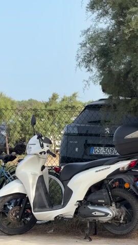 A white scooter is parked in front of a fence. A black SUV is parked behind the scooter. Trees and bushes are in the background. The sky is clear and blue.