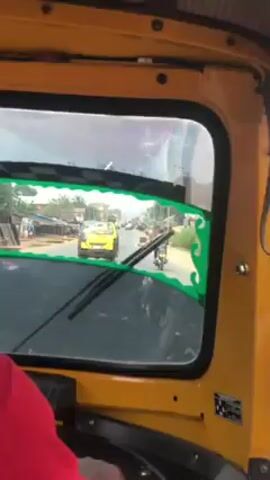 The video, taken from the inside of a yellow vehicle with a window decorated with green accents, shows a view of a road with several vehicles, including another yellow vehicle and a motorcycle. The road appears to be in a town or city, with buildings visible in the background. The camera seems to be moving slowly, as if the vehicle is in traffic. A person wearing a red shirt is partially visible in the foreground.
