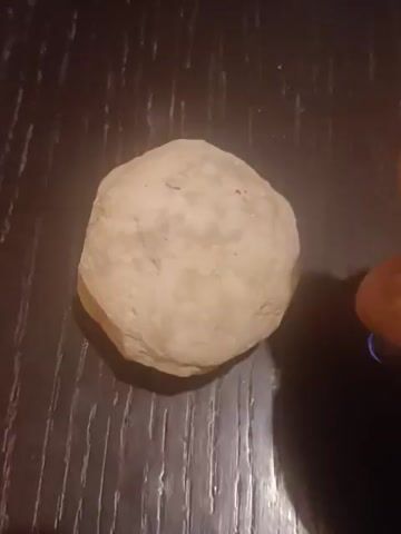 A close-up shot focuses on a light-colored, smooth, oval-shaped object, possibly a stone or mineral, resting on a dark, textured surface resembling wood grain. A bright white light, emanating from a small, cylindrical device held by an unseen hand, illuminates one side of the object, causing it to glow. The light source is positioned to the right of the object and casts a soft glow and shadow. The scene is dimly lit, with the focus entirely on the illuminated object.