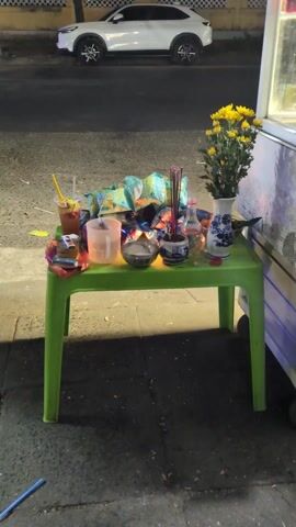 The video shows a small, bright green plastic table on a sidewalk. On the table is a small altar set up, with various items arranged on it. To the left is a glass of fruit drink with a straw, and a clear plastic pitcher filled with water. Next to those is a small silver bowl containing rice. Adjacent to the rice bowl is a small blue and white ceramic pot filled with incense sticks, some of which are lit and emitting smoke. There's also a small glass bottle, a small shot glass, and a partially covered red cap. A white and blue patterned vase with bright yellow flowers is on the right side. Behind those objects are several packages of snacks, piled in a heap. In the background, a white car is parked on the street. The overall lighting suggests it is evening or nighttime.