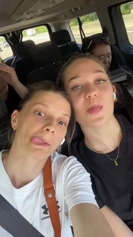The video shows two young women taking a selfie in the back seat of a car. They are both wearing casual clothing and appear to be smiling. The woman on the left is wearing a white t-shirt with a red strap bag over her shoulder. The woman on the right is wearing a black t-shirt. In the background, other passengers can be seen in the car, as well as the road outside.