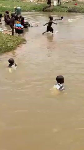 A short video shows several young children playing and swimming in a shallow, muddy river. On the left bank, several people are doing laundry in tubs. Some children are also standing near the bank, watching or preparing to enter the water. The river is surrounded by grassy vegetation and scattered debris. The overall atmosphere appears carefree and playful.