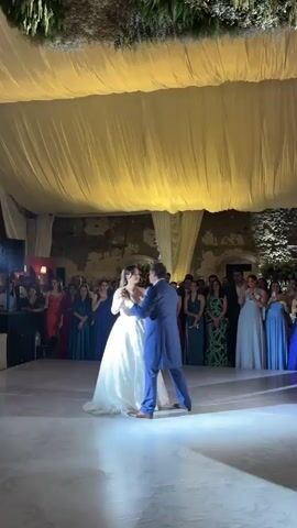 A newlywed couple is dancing at their wedding reception. The bride is wearing a white, strapless wedding gown with a full skirt, and the groom is in a blue suit. They are surrounded by wedding guests, who are standing and watching them dance. The reception is being held in a large room with a white fabric ceiling and weathered stone walls. The room is lit by warm, yellow lighting.