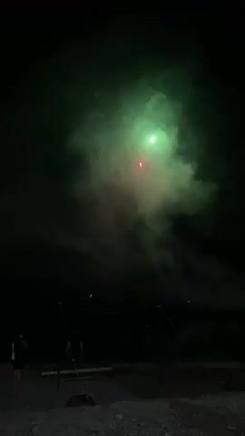 The video shows a night sky with fireworks. In the first frame, yellow and red fireworks are visible amidst smoke. The second frame displays small dots of light, possibly distant fireworks, against a dark background. The third frame shows a green firework explosion with smoke. The fourth frame features a red firework with accompanying smoke. The final frame shows a completely dark sky with some faint smoke remaining. In the foreground, silhouetted figures of people are present, along with some poles and a bench. The ground appears to be rocky or sandy.