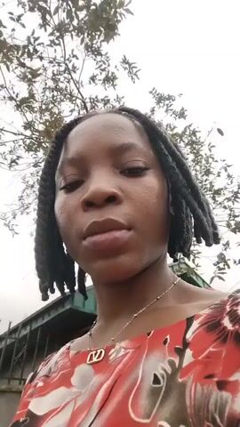 A young woman with dark braided hair is looking into the camera. She is wearing a red and white floral print top and a silver necklace. The background appears to be outdoors with trees and a cloudy sky. The video is short and shows the woman's face from the chest up. She blinks a few times and turns her head slightly.