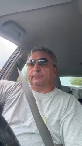 A man with short, graying hair and a light gray t-shirt is sitting in the driver's seat of a car, wearing aviator sunglasses. He is looking forward, and a seatbelt is across his chest. The interior of the car is visible, including the ceiling and the side window. Outside the car, greenery and a cloudy sky can be seen.