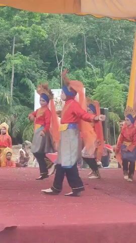 This is a video of a group of children performing a traditional dance outdoors. They are on a red stage, with a yellow canopy overhead. Behind them is a backdrop with images of people and text in a foreign language. The children are dressed in colorful, ornate costumes, including red shirts, patterned skirts, and headscarves with orange veils. They are all wearing similar headdresses with golden accents. The dancers are moving in unison, with their arms outstretched and their bodies in various poses. The backdrop suggests that this is a cultural event or festival. The background shows a lush green forest and trees, indicating an outdoor setting. Some audience members can be seen in the background, seated or standing. The lighting suggests it is daytime.