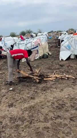 This video captures a scene in a refugee camp. A person wearing a red t-shirt and light-colored pants is bent over, working with a pile of sticks and branches on the muddy ground. The individual appears to be preparing firewood.

In the background, numerous temporary shelters made of white tarpaulins and other materials are visible. Many of these shelters are wrapped with ropes and straps, and some have the United Nations High Commissioner for Refugees (UNHCR) logo printed on them, indicating UNHCR assistance. There are also large, bundled items, possibly supplies or more building materials, stacked near the shelters.

Other people can be seen in the distance, some near the shelters and others in the background, suggesting a populated settlement. The ground is uneven and appears to be made of dark, damp soil, with some debris scattered around. The sky is overcast, suggesting a cloudy day. The overall setting is indicative of a temporary living situation, likely a displacement camp, given the nature of the shelters and the presence of UNHCR branding. The activity of collecting and preparing firewood is a common and essential task for survival in such environments. No discernible text other than the UNHCR logo is visible.