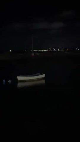 The video displays a nighttime scene of a calm body of water with several boats anchored. The water reflects the faint lights from a distant shore, where buildings and a tall pole are visible. The camera pans slowly across the water, revealing two small boats in the foreground and a larger boat further in the distance. The atmosphere is dark and quiet.
