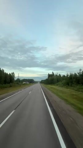 The video shows a long, paved road with white lines marking the lanes. The road is flanked by green grass and trees on both sides. The sky is cloudy and the lighting suggests it is either early morning or late evening. There are no other vehicles or people visible in the video. The camera appears to be mounted on a moving vehicle, likely a car, as it captures the perspective of someone traveling down the road. The overall scene conveys a sense of travel and open space.