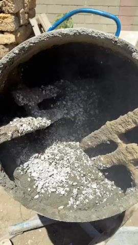 A concrete mixer is shown from the inside, with the drum rotating. The mixer contains cement, gravel, and water. The concrete is being mixed by the paddles inside the drum. In the background, there are stacks of bricks and a wall. There is also a blue handle for the mixer and a red glove on the ground.