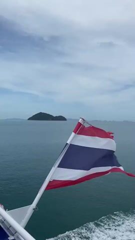 The video shows a ferry sailing on a body of water, possibly the ocean. In the beginning, the Thailand flag is shown waving in the wind. As the camera pans, a life raft and several rows of empty white benches on a blue deck are visible. Later, several people are shown relaxing and enjoying the view from the deck. One person is sitting, another is touching her hair, and a shirtless man is leaning on the railing, looking out at the land in the distance. In the background, there is a cloudy blue sky, islands, and what appears to be a shoreline with land and mountains.