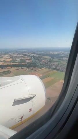 This video shows a view from an airplane window as it flies over a rural landscape under a clear blue sky. In the foreground, a large white airplane engine with yellow accents is visible, with some text and warning symbols on its housing. Below the plane, a winding river flows through agricultural fields, and a small town with red-tiled roofs is situated along its banks. The fields are a mix of green and brown, suggesting agricultural activity. In the distance, the landscape appears flatter and hazier. The time of day appears to be daytime, with bright sunlight. The weather seems clear and sunny. The scene evokes a sense of travel and distant observation, capturing a moment of flight over a picturesque European countryside.

The visible text on the engine housing includes:
- A red circle with a diagonal line through it, over a symbol resembling a flame or exhaust, indicating no fire or no exhaust in that area.
- Text below that is partially obscured, but seems to include an international symbol of "No Smoking" or "No Open Flames" and possibly other safety instructions.
- The word "RYANAIR" is visible in yellow lettering on a black background along the bottom edge of the engine housing.