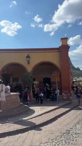The video shows a sunlit public square. Several people are standing around a stone fountain in the center, some interacting with it. There are buildings around the square, with architectural features like arches and balconies. The sky is blue with scattered clouds. The square is paved with cobblestone. Some people are walking through the square, seemingly tourists. The lighting suggests it is daytime. There are shops and businesses lining the buildings.