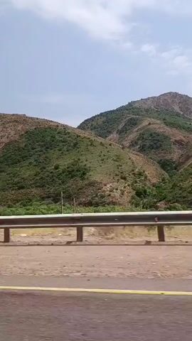 The video shows a scenic view of a mountainous landscape from a moving vehicle. The mountains are covered in green vegetation, with some bare rocky areas visible. A guardrail runs along the side of the road in the foreground, with a yellow line painted on the asphalt. The sky is partly cloudy.
