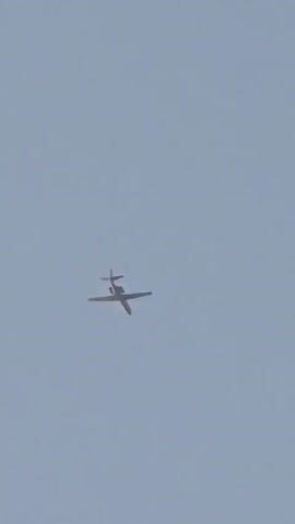 A small, twin-engine aircraft is shown flying against a pale blue sky. The aircraft appears to be a turboprop or jet, with its wings extended horizontally and its tail fins visible. The image is static, with no movement detected in the video. The angle of the camera suggests it is looking up at the aircraft from the ground. No people or other objects are visible in the frame. The lighting is consistent with daylight, but the sun's position is not discernible.