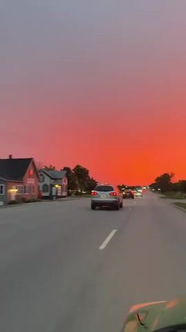 This vertical video captures a car driving on a road during a vibrant sunset. The sky is a dramatic gradient of colors, transitioning from a soft grey-blue at the top to a fiery red-orange near the horizon. The setting appears to be suburban or semi-rural, with trees and some buildings lining the road.

Several cars are visible on the road, some in motion and some appearing to be parked or stopped. The perspective suggests the video is filmed from within a vehicle, looking forward. The motion blur indicates the car is moving at a moderate pace. The time of day is clearly sunset, characterized by the intense coloration of the sky. There are no people clearly visible, but their presence is implied by the vehicles and the setting. The overall mood is one of serene beauty, amplified by the dramatic natural light display. No text is visible in the video. The location context indicates this scene is taking place in Carol Stream, United States of America.