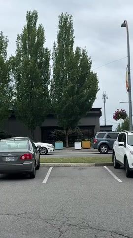 The video shows a parking lot with several cars and a motorcycle. In the foreground, a dark gray sedan is parked in a spot. To its right, a white sedan is parked. Further back, a white pickup truck and a blue SUV are visible. Behind these vehicles, there is a row of tall trees. In the background, there is a building with dark siding and large windows. A cell tower is visible on the right side of the frame. A bus stop shelter with advertisements is also present. A blue motorcycle is parked between a gray SUV and a white car, with a person wearing a helmet sitting on it. The sky is overcast.