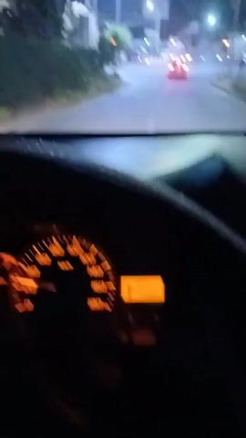 The video captures a nighttime view from inside a vehicle looking out at a street intersection. Streetlights illuminate the scene, casting a glow on the asphalt road and surrounding buildings. A red car is visible in the distance, moving away from the camera and turning right. Other vehicles and streetlights are present, creating a typical urban nighttime environment. The interior of the car, including the dashboard, is partially visible in the foreground. The overall impression is one of a brief journey through a well-lit street at night.
