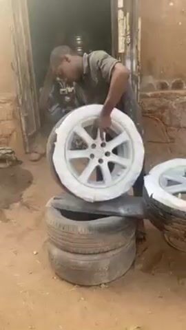 A man is spray painting the rims of car tires. He is standing in front of an open doorway of a building with a dirt yard. He is wearing a dark t-shirt and pants, and he has a paint spot on his shirt. The man is holding a spray paint gun in his right hand and using his left hand to hold the wheel steady. He is focused on the task at hand. The tire that he is working on is placed on top of a stack of old tires. Another tire with its rim painted is sitting beside the man on an old tire. The car rims are white. The environment is dusty and cluttered.