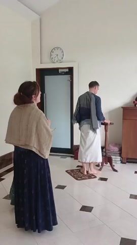 Here's a detailed description of the video:

The video shows an indoor scene with a group of people in what appears to be a temple or religious gathering space. The room has white walls and a tiled floor with black square accents.

At the start, a woman in a long blue skirt and a beige shawl is standing near a doorway. A man wearing a white dhoti and a blue shirt with a jacket draped over his shoulders is walking toward a small wooden table. 

As the camera shifts, two bald men wearing dhotis and one wearing a maroon hooded sweatshirt enter the frame. One man is playing a drum, which looks like a mridanga or similar percussion instrument. They are walking towards a large glass door, which is likely the entrance. Through the glass door, a glimpse of an outdoor area with trees and grass is visible.

The men seem to be participating in some sort of ritual or procession with music. The room also has a wooden cabinet near the wall. A clock is visible on the wall, reading around 11:10. A cross is also displayed.