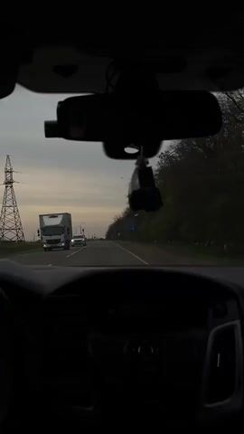 The video captures a first-person perspective from inside a car driving on a road during what appears to be dusk or early evening, characterized by an overcast sky. The dashboard, steering wheel, and a portion of the driver's hand are visible in the foreground. A rearview mirror with a dangling air freshener or charm and a dashcam mounted below it are prominent. Heart-shaped cutouts or stickers are seen on the upper windshield area.

The road ahead is a two-lane asphalt surface. On the right side of the road, trees and dense foliage line the path. On the left, utility poles with power lines are visible, along with streetlights further down the road. Several vehicles are seen on the road: a large white truck is approaching on the left, and a white car with headlights on is also visible in the distance.

A blue road sign with white text is present on the right side of the road. The top line of text clearly reads "СТАРОМИНСКАЯ" (Starominskaya), with additional text and an arrow below it, indicating a turn. This sign provides a geographical cue, consistent with the location context of Pervomayskiy, Russia.