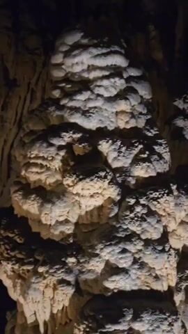 A panning shot of a cave interior. The cave is filled with stalactites and stalagmites, illuminated by spotlights. A winding staircase with railings and lights is visible on the right side of the frame, leading down into the cave. The rock formations are rough and textured, with some appearing smooth and others more jagged. The lighting creates shadows and highlights, emphasizing the depth and unevenness of the cave walls and formations.
