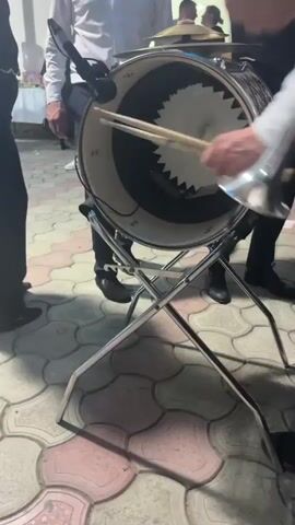 A close-up shot focuses on the inside of a bass drum, which rests on a chrome stand. A person's hands are visible, adjusting something inside the drum. Several people stand around the drum, their legs and lower bodies visible. The floor is paved with interlocking stones in a repeating pattern of light pink and beige. The lighting suggests an outdoor setting, possibly at dusk or during an event.