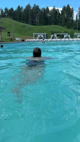 A person with dark hair in a bun swims in a blue swimming pool, facing away from the camera. The pool is surrounded by lounge chairs and cabanas, with trees and a grassy hill in the background. The sky is blue with some clouds.