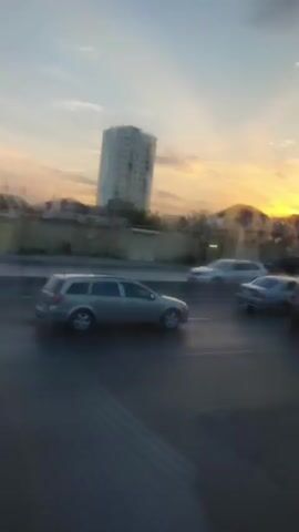 A blurred video shows a street scene at dusk. Several cars are visible on the road, some in motion. In the background, a tall, light-colored building stands out against the sky, which has a soft, hazy glow. The overall impression is one of a typical urban environment during twilight.