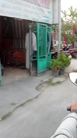 The video shows a first-person view of someone riding a motorcycle through a street. The street appears to be in a residential or semi-commercial area. There are several motorcycles parked on the side of the road. Buildings with red and green accents are visible, along with gates and trees. In the background, there are small food stalls or kiosks. Some people are seen near the stalls. There are flags and banners displayed. The road surface is rough and cracked, with puddles of water visible, suggesting it might have rained recently. The lighting suggests it is daytime, possibly late afternoon.