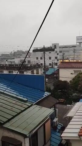 A static, eye-level medium shot captures a view of a town under an overcast sky, likely during or just after rain, as indicated by the wet appearance of the tiled roofs. In the foreground, several houses with different colored roofs are visible: a blue roof, a green roof, and an orange tiled roof. A black power line stretches diagonally across the upper portion of the frame, originating from the top left and extending towards the center right. In the middle ground, more houses and buildings are seen, including a white building with a red roof and a larger white building in the background with a sign that reads "S&D". Trees with dark green foliage are scattered throughout the scene, and utility poles with wires are present. The overall impression is one of a quiet, residential area experiencing gloomy weather.