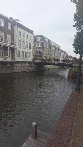 The video pans from right to left, showing a canal with buildings lining its banks. The buildings are multi-story structures with a variety of architectural styles, some appearing older with red brick accents and others more modern. The canal water is dark and rippling, reflecting the overcast sky. In the foreground, there is a wooden dock or walkway. Further down the canal, a small boat is visible, and a lamppost stands on the brick pathway beside the canal. The scene appears to be in a European city, likely with a historical canal system. There are no people visible in the video.