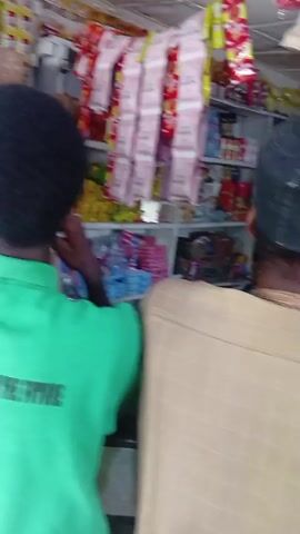 Inside a small store, a man in a green t-shirt with writing on the back stands at a counter, another man in a yellow and maroon t-shirt looks at shelves of goods. The shelves are filled with packaged food, drinks, and various items. A third man wearing a tan shirt with lines stands near the counter. People seem to be shopping or waiting to be served in the store.