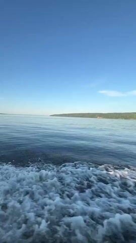 This media file captures a boat ride on a calm body of water, likely a lake or a bay, in Siukterka, Russia. The scene is dominated by a vast expanse of blue sky and tranquil water, suggesting fair weather and a peaceful atmosphere. The time of day appears to be late afternoon or early evening, indicated by the low angle of the sun, which casts a warm, golden glow on the water and creates a shimmering reflection.

The primary subject is the boat itself, seen from a low-angle perspective looking forward. We see parts of the boat's interior, including a wooden deck in the foreground and a grey hull. A pair of work gloves, one white and one green, are resting on the deck, hinting at recent activity or preparation. The boat is in motion, evidenced by the frothy white wake trailing behind it.

The background consists of a distant, tree-lined shore, adding a natural element to the serene landscape. The horizon is sharp and clear, with no visible signs of pollution or significant human development on the distant landmass. The overall mood is one of tranquility and gentle movement, evoking a sense of escape and connection with nature. The clear blue sky and the smooth water contribute to a feeling of calm and serenity. There is no visible text in the media file.