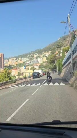 The video shows a view from inside a vehicle, looking out through the windshield. The vehicle is driving on a paved road with clear white lane markings and a black and white striped speed bump. The road curves to the right as it ascends a hill. On the left side of the road, there is a retaining wall, trees, and buildings. On the right side of the road, there is a steep hillside covered in vegetation and some construction.

In the distance, a city with many tall buildings is visible, nestled on a hillside overlooking the ocean. The sky is clear and blue. Several vehicles are on the road: a black van, a white van, a red car, and a motorcycle with a rider wearing a helmet and black clothing. The motorcycle is in front of the black van. The camera is angled slightly downwards, showing the dashboard of the vehicle, including a phone mounted on a holder. The overall impression is of a sunny day driving through a scenic, urban environment on a coastal hillside.