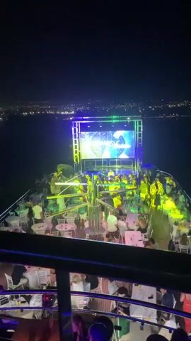 This is a night-time video taken from the deck of a boat at sea. A large screen displaying a logo with the text "OCEANO CLUB" is visible at the front of the boat, illuminated by stage lights. Below the screen, a crowd of people are gathered, with some seated at tables and others standing. The boat is moving, as evidenced by the water's surface. In the background, a distant city skyline is visible with numerous lights. The overall atmosphere appears to be that of a party or club event on a boat.