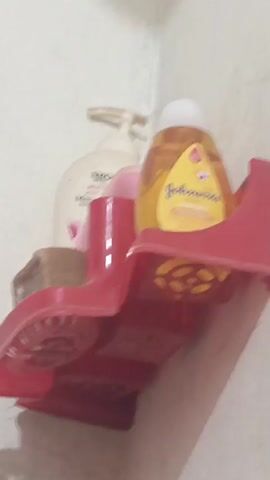 The image presents a close-up view of a red plastic shower caddy mounted on a plain, light-colored wall, likely within a bathroom or shower area in Kakki, Pakistan. The caddy is positioned at a slightly low angle, looking upwards.

On the right side of the caddy, a yellow plastic bottle of Johnson's baby shampoo is prominently displayed. Its white cap is visible at the top, and the yellow label clearly shows the "Johnson's" brand name and "baby shampoo" text. The bottom of the caddy beneath this bottle has circular, slotted cutouts, presumably for drainage.

To the left of the Johnson's bottle, a partially visible brown bar of soap rests on the caddy. Behind the soap and further to the left, a white pump-top bottle is present. Its label shows parts of the text "SMOOTH," "BODY," and "Moisturizing." A small, mostly obscured pink bottle is tucked behind the soap and the white pump bottle.

The wall behind the caddy is uniform in color, providing a simple background to the toiletries. There are no people visible, and the scene depicts a static arrangement of personal hygiene products. The lighting appears to be indoor and even, with no strong shadows indicating a particular time of day or outdoor conditions.