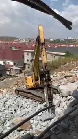 The video shows a yellow excavator with a jackhammer attachment breaking up large rocks on a construction site. The excavator is positioned on a pile of broken rocks, with a residential area visible in the background. The sky is bright blue with some clouds. The video is taken from an elevated position, with some bamboo supports visible in the foreground. The excavator arm is up and then comes down with the jackhammer to smash another rock.
