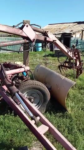 The video shows a rural scene with farm equipment and a chicken. In the foreground, a red mechanical arm extends towards the viewer, connected to a wheel and part of a larger machine. To the right of the arm, a large, cylindrical object, possibly a roller, lies on the ground. Behind it, a chicken with brown and white feathers pecks at the grass. Further in the background, a dilapidated building with a corrugated metal roof and a wooden fence is visible under a clear blue sky. The ground is covered in green grass and some scattered debris. The video is shaky, suggesting it was filmed by someone holding a camera or phone.