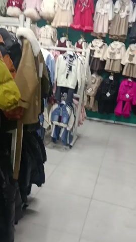 The video depicts a shopping mall or department store setting. Various clothing racks are visible, showcasing items such as jackets, shirts, and pants. Customers can be seen browsing through the merchandise. One person is mopping the floor. The lighting appears bright and artificial.