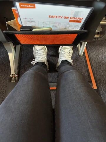 The image is captured from a first-person perspective, looking down at the legs and feet of a person seated in an airplane. In front, the back of an airplane seat with an integrated pocket is visible.

The scene depicts someone seated inside an airplane cabin, likely either boarding or during a flight.

The person's legs are clad in dark grey or black skinny jeans, and they are wearing white sneakers with white laces. The seat directly in front features a black backrest with an orange mesh seatback pocket. A white "SAFETY ON BOARD" card, bearing the easyJet logo, is prominently displayed in the pocket, along with what appears to be a small black electronic device (possibly a phone) and a pen or stylus. The floor beneath the feet is covered with a dark grey, subtly patterned carpet. The cabin appears brightly and evenly lit, suggesting artificial lighting. No external weather conditions or time of day are discernible. The shot emphasizes the limited legroom typical in economy airline seating.

The text "easyJet" and "SAFETY ON BOARD" are clearly visible on the information card in the seat pocket.