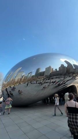 The video shows the Cloud Gate sculpture in Chicago, also known as “The Bean”, with a crowd of people around it. The sculpture’s reflective surface mirrors the Chicago skyline and the people nearby. Individuals are walking around, taking photos, and interacting with the artwork. The weather appears sunny with a blue sky and scattered clouds. Tall buildings can be seen in the background, framing the scene. The area surrounding the sculpture is paved with light gray tiles.