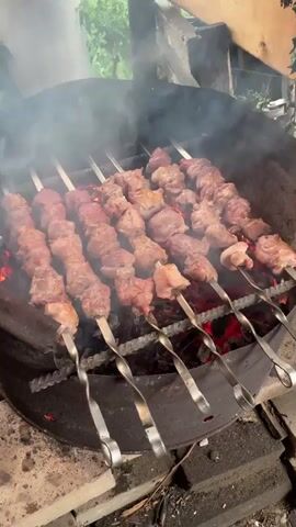 This is a close-up, eye-level shot of several skewers of meat cooking over hot coals. The meat appears to be beef or lamb, cut into bite-sized pieces and threaded onto metal skewers. Smoke rises from the grill, obscuring some of the meat. The grill itself is a large, round metal basin with a wire rack supporting the skewers. The background is blurred, but suggests an outdoor setting with greenery and wooden structures. The overall impression is one of a barbecue in progress.