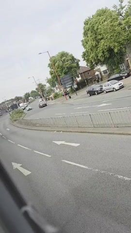 This media file captures a street view from a moving vehicle, likely a bus, in Birmingham, United Kingdom. The scene is a moderately busy road with multiple lanes and directional arrows painted on the asphalt. Several cars are visible on the road, including a red sports car, a white hatchback, and a dark-colored SUV. The surrounding environment features residential houses with pitched roofs and lush green trees, suggesting a suburban or urban fringe setting. Streetlights and utility poles line the road, indicating infrastructure typical of a developed area.

The sky is overcast, suggesting a cloudy or temperate day, with no strong indication of the time of day beyond it being daytime. The overall atmosphere appears calm and ordinary, depicting a typical urban commute. There is a metal fence separating the road from a grassy verge. Some people are visible walking on the sidewalks or near the houses. While no specific event is taking place, the footage documents the everyday flow of traffic and the urban landscape. No discernible text is visible in the media file.