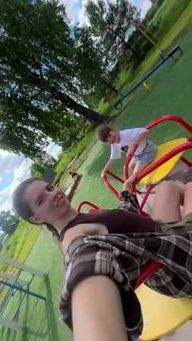A young woman and a child ride a yellow and red merry-go-round on a playground on a bright, partly cloudy day. The woman is wearing a brown tank top, green shorts, and a brown and black plaid shirt. She has her hair pulled back in a ponytail. The child is wearing a white t-shirt and blue shorts. The woman is holding a phone and filming. The playground has green artificial turf and various play structures. Buildings and trees are visible in the background. The woman and the child are both smiling and appear to be enjoying themselves.