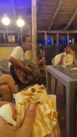 The video shows a partially eaten wrap being held by a person, with other people visible in the background. The wrap is in a yellow and white striped paper sleeve, filled with what appears to be lettuce and meat. The person is sitting at a table. A man is playing an acoustic guitar and singing in the background. Another person is sitting at a table, eating. The video is taken at a juice bar, which is indicated by a blue neon sign. The scene takes place in an outdoor setting with a thatched roof and exposed wooden supports. String lights are visible in the background.